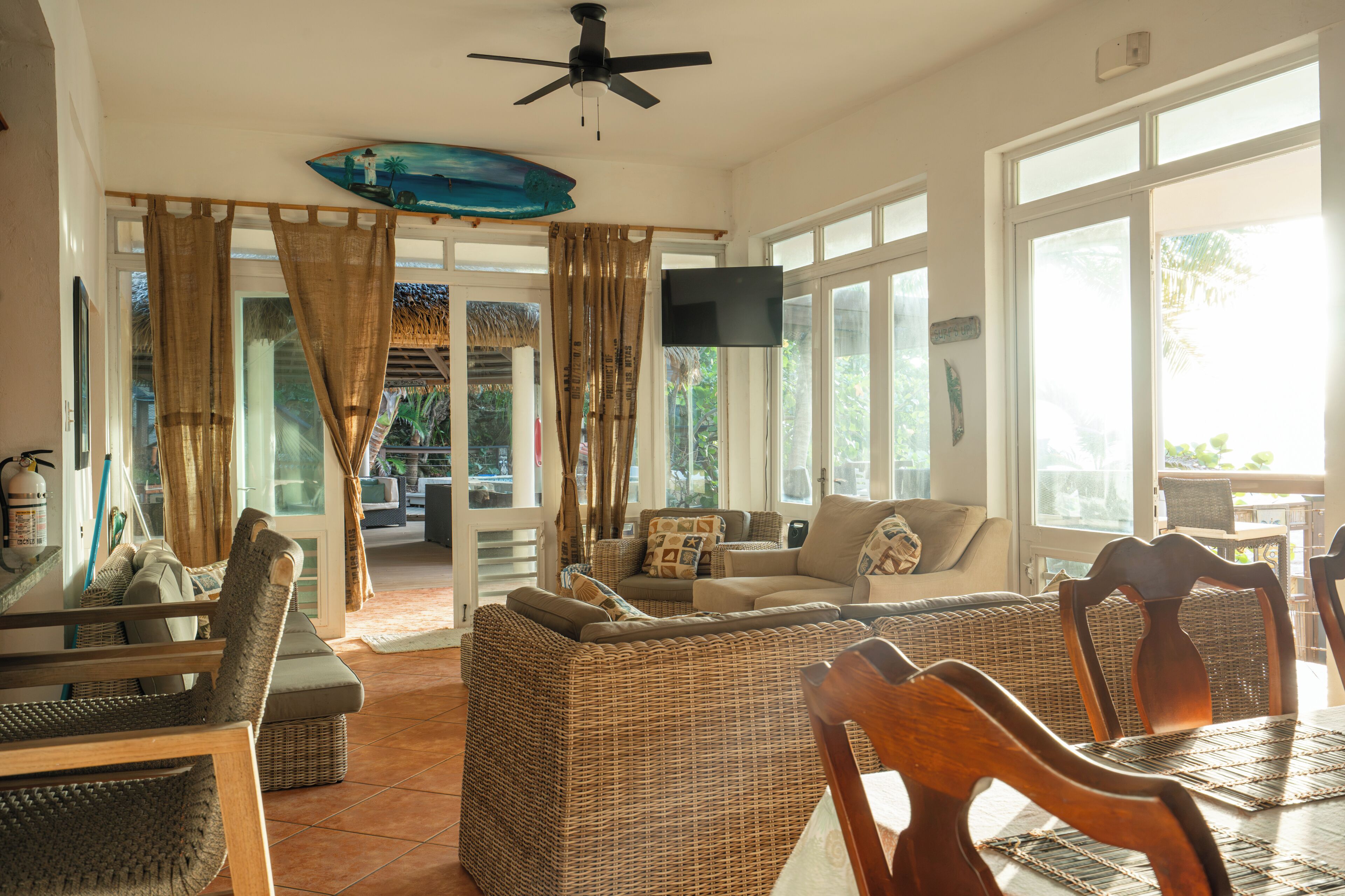 A spacious living area with a large sofa, dining table, and chairs, a ceiling fan, and a TV mounted on the wall.