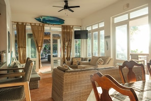 A spacious living area with a large sofa, dining table, and chairs, a ceiling fan, and a TV mounted on the wall.