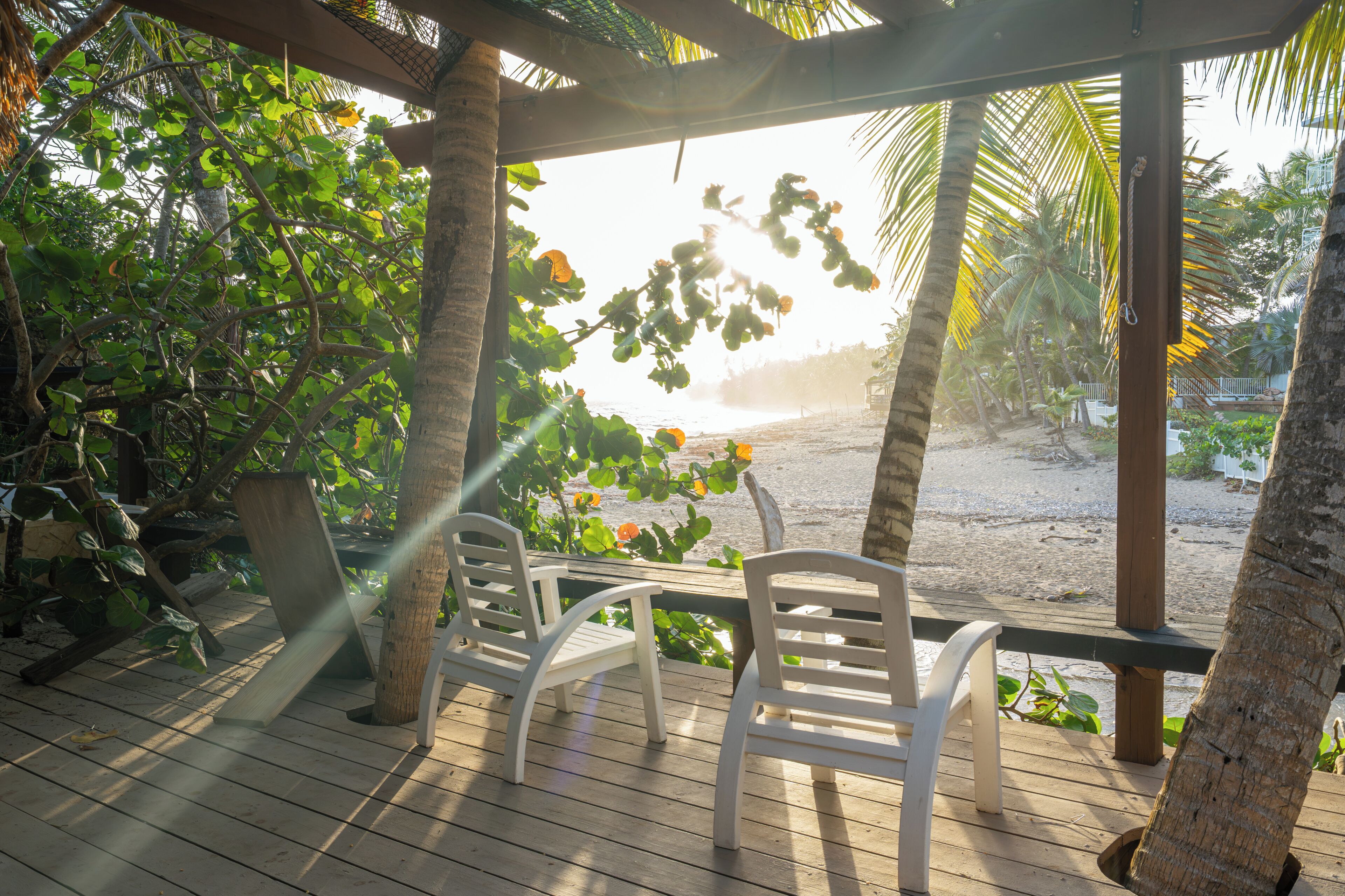 A wooden deck with two white chairs, overlooking a beach with palm trees and a clear view of the ocean.