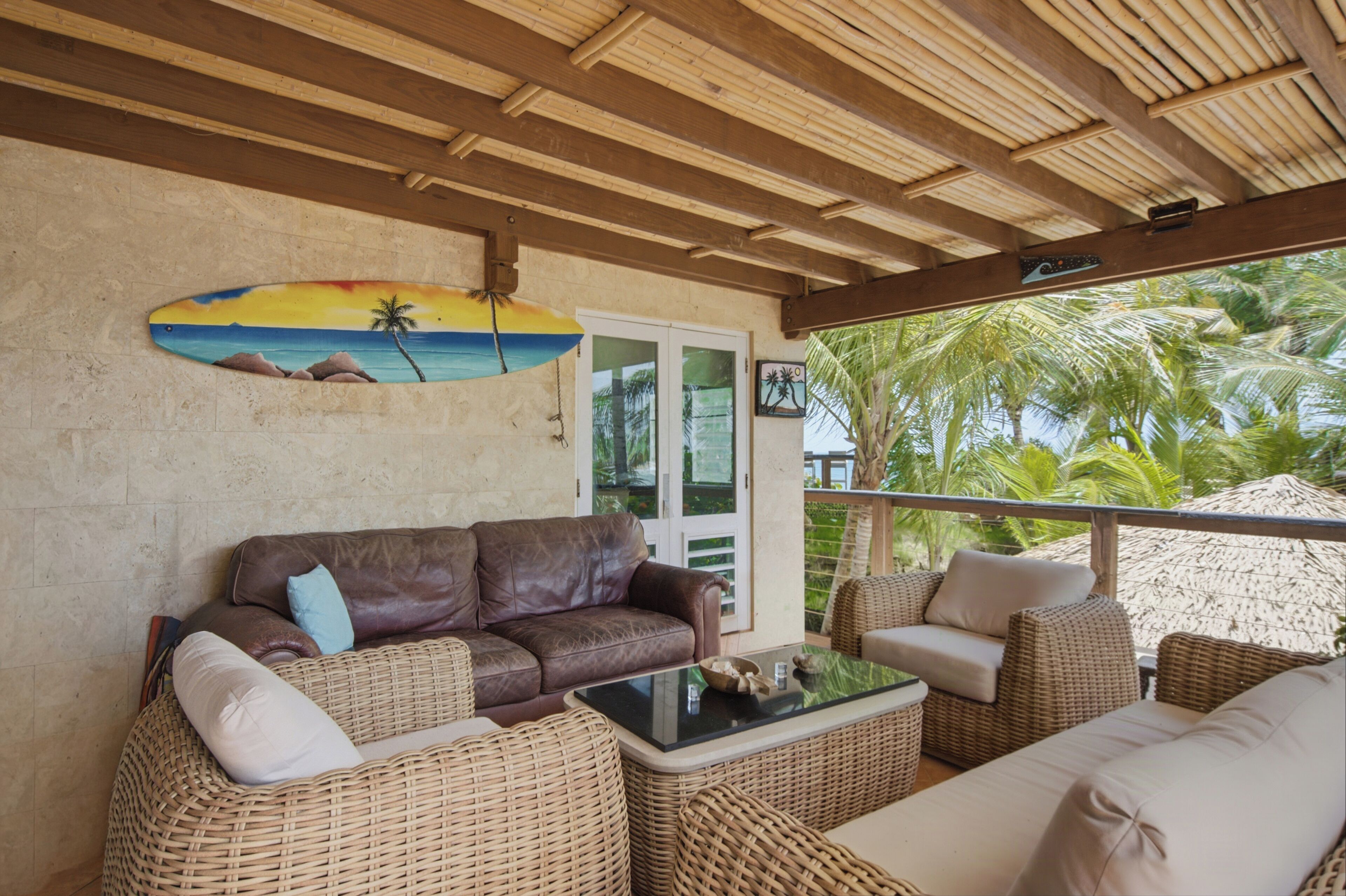 A covered outdoor seating area with a brown leather sofa, wicker furniture, and a glass table.