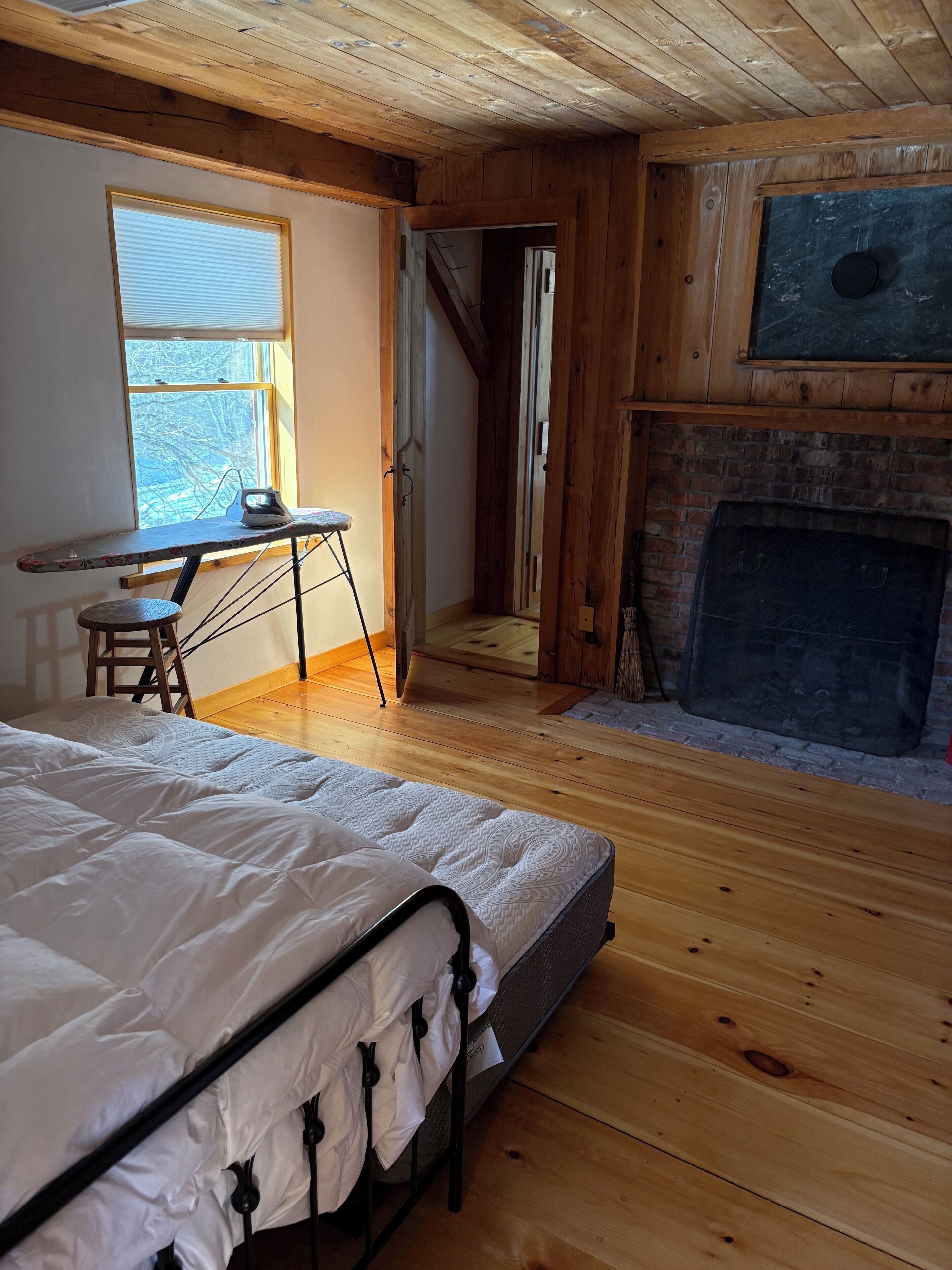A bedroom with a bed, a desk with an iron, a fireplace, and a window with blinds.
