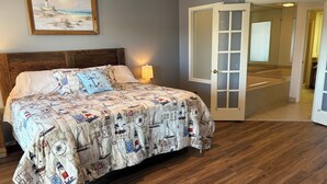 Et soveværelse med sengegavl i træ, en seng med quilt i maritimt tema, et natbord med lampe, og en dør til et badeværelse med badekar.