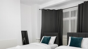 Una camera d'albergo con due letti, un lampadario, un'ampia finestra con tende e un quadro appeso al muro.