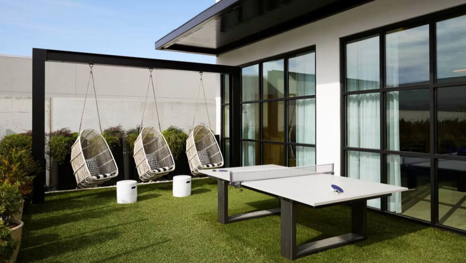 Un espace extérieur moderne comprenant une table de ping-pong, trois fauteuils suspendus et un petit jardin.