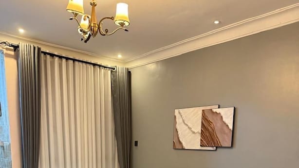 A modern living room with a grey sofa, two wooden coffee tables, a chandelier, and a large window with curtains. - Clover Homes Ug (Kampala)