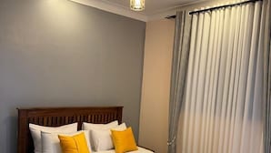 A bedroom with a wooden bed, white and yellow pillows, a nightstand, and a ceiling light. - Clover Homes Ug (Kampala)