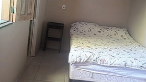 Una habitación pequeña y tenuemente iluminada con una cama individual, una mesita y una ventana con la puerta entreabierta.