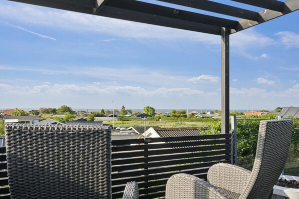 Een overdekt terras met rieten meubilair, een pergola en uitzicht op huizen en groen.