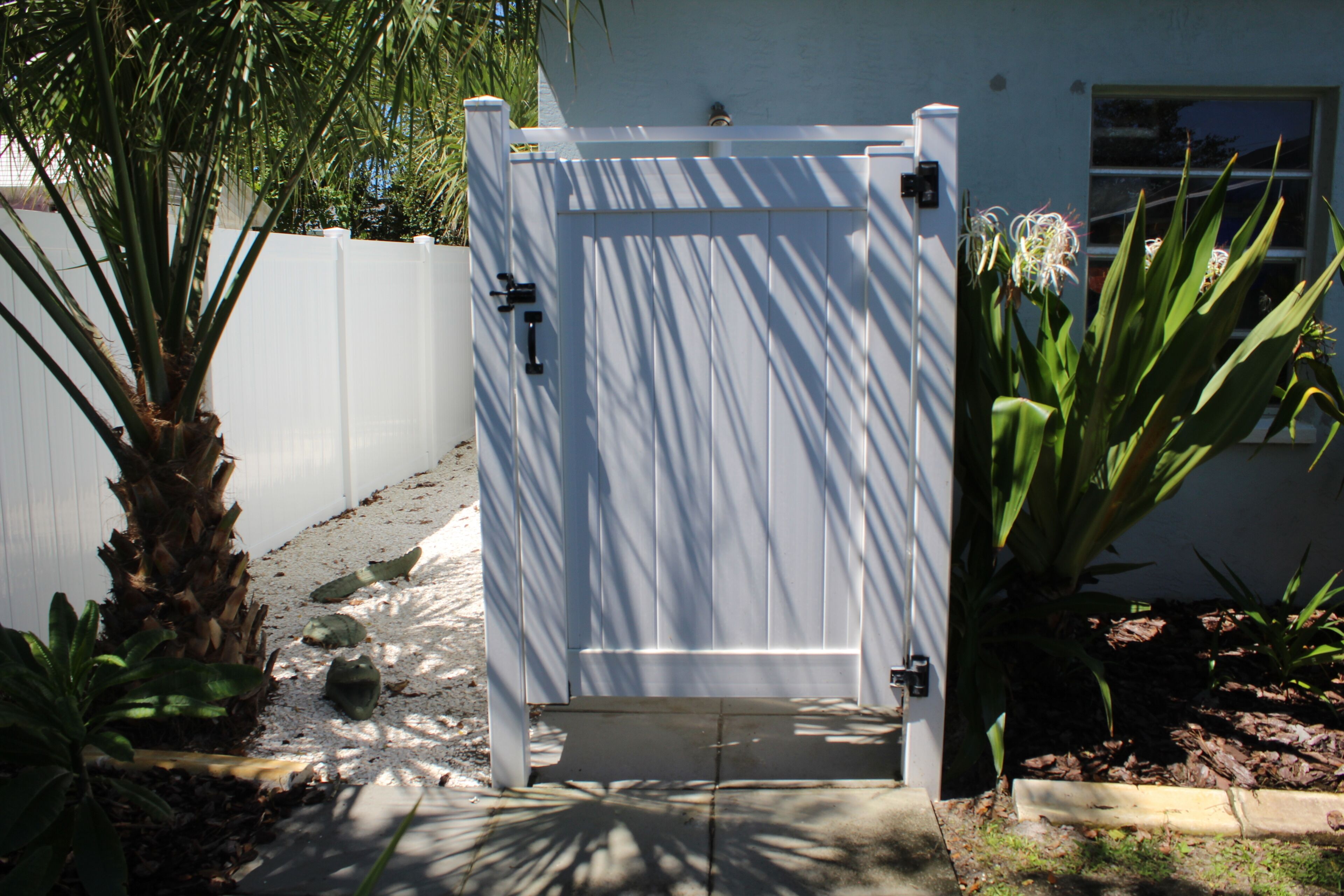 A gated entrance with a white fence, a palm tree, and a plant with long leaves.