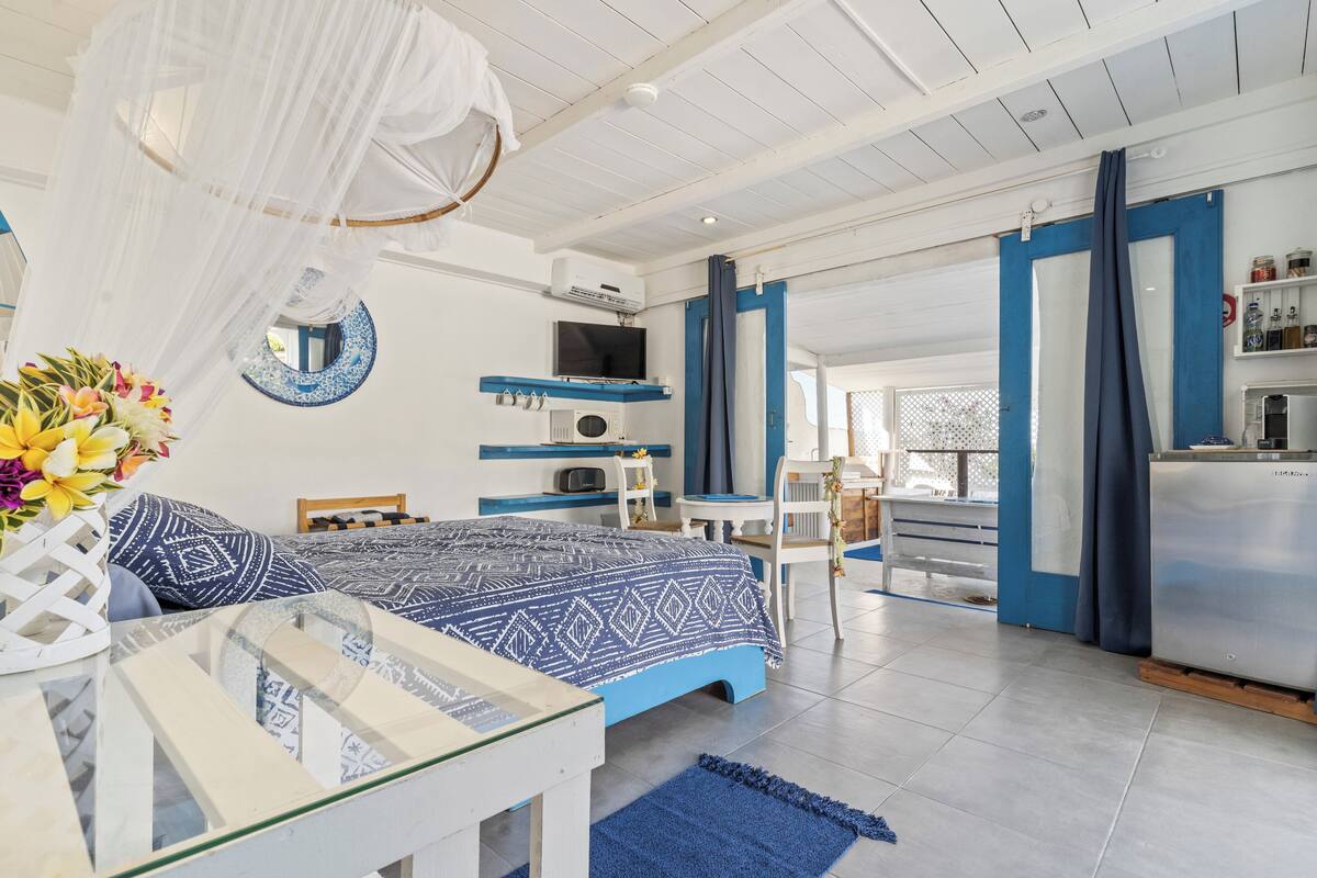Een moderne slaapkamer met een bed, nachtkastje en spiegel. Er is een kitchenette met een koelkast en fornuis.
