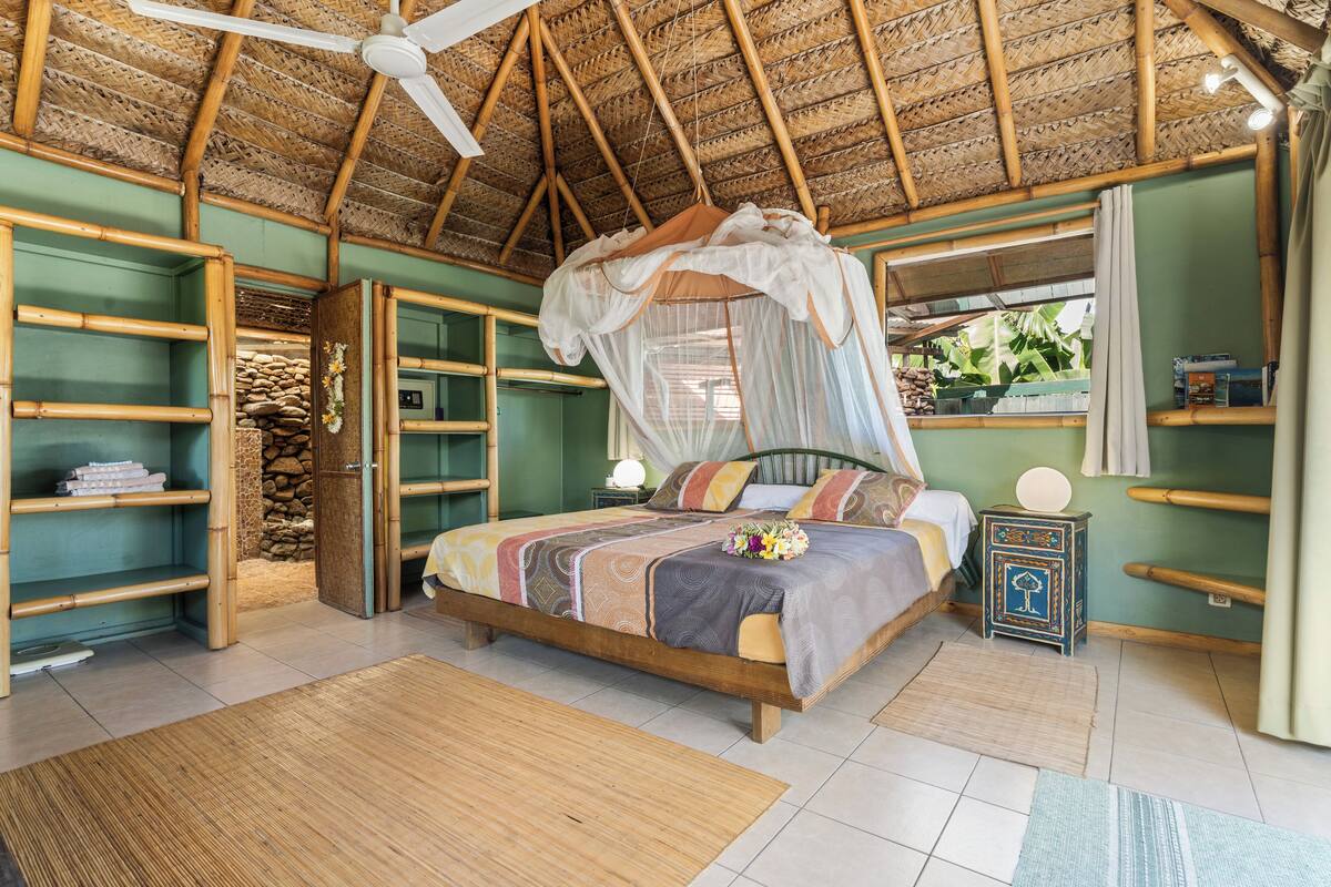 Een kamer met een bed, een plafondventilator, een nachtkastje en een raam met uitzicht op groen.