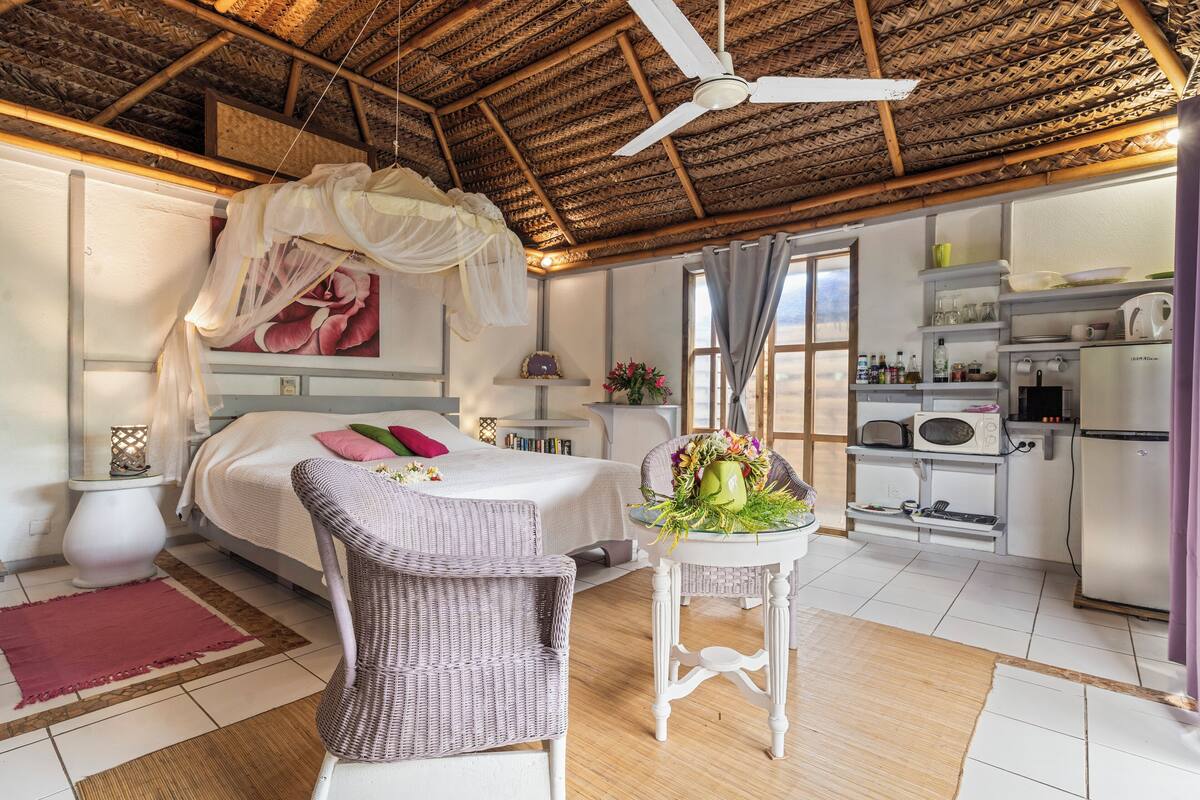 Een slaapkamer met een bed, een rieten stoel, een nachtkastje met een bloevas en een plafondventilator.
