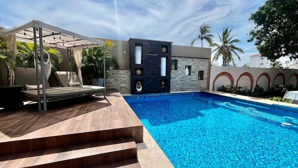 A modern outdoor pool area with a stone wall, a canopy, and a set of stairs leading to a lounge area.