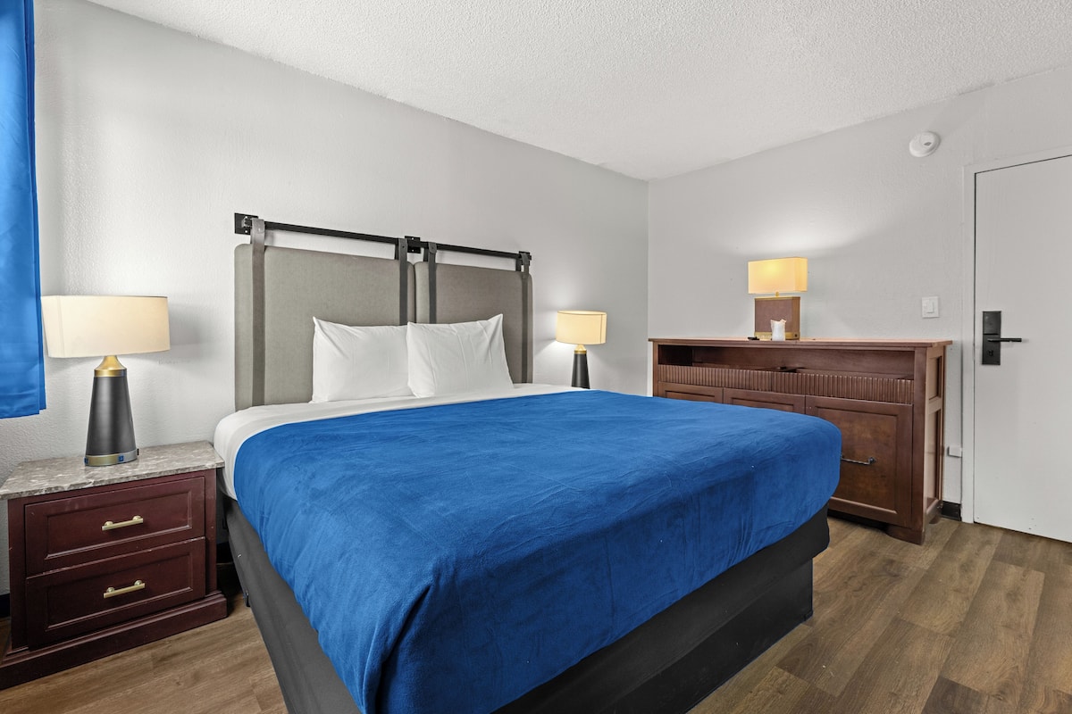 Una habitación de hotel con una cama, dos mesitas de noche con lámparas, un aparador de madera y una puerta.