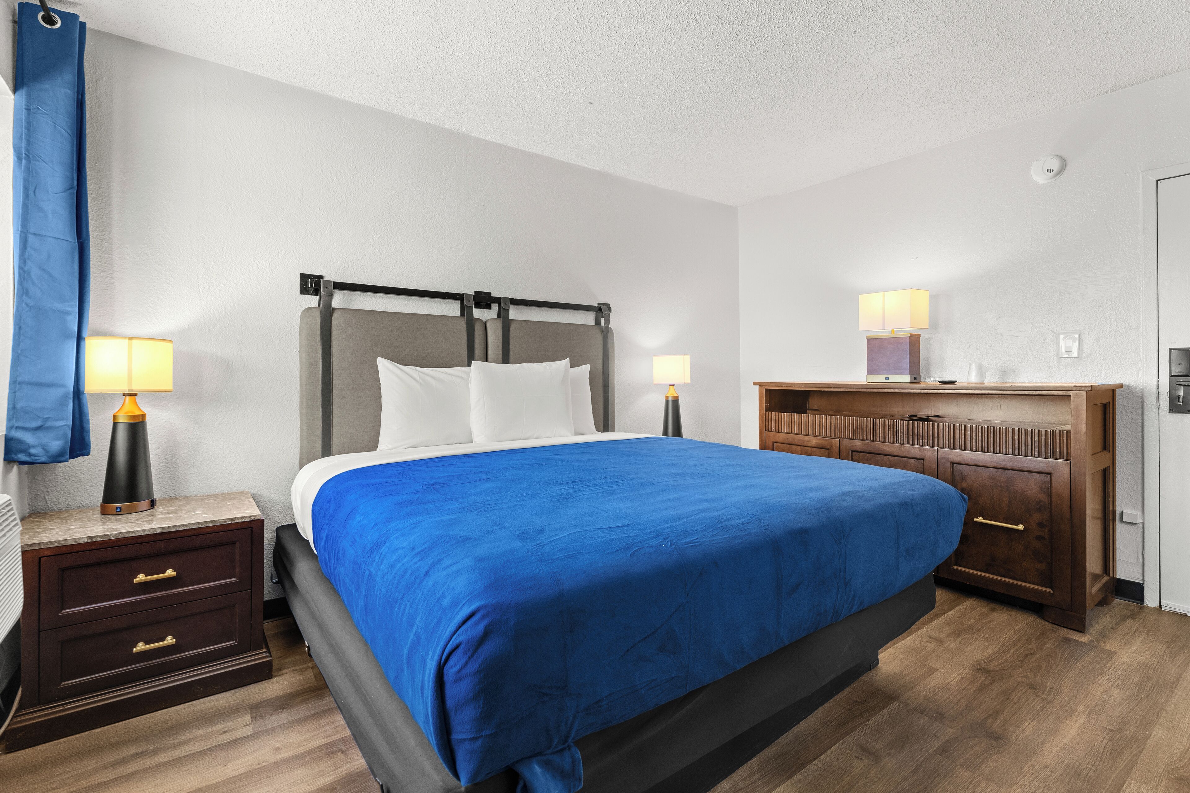 A hotel room with a bed, nightstands, lamps, a wooden dresser, and a door.