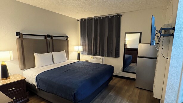 A hotel room with a bed, bedside table, lamp, air conditioning unit, and a refrigerator. - Studio Lodge Hotel (North Hollywood)