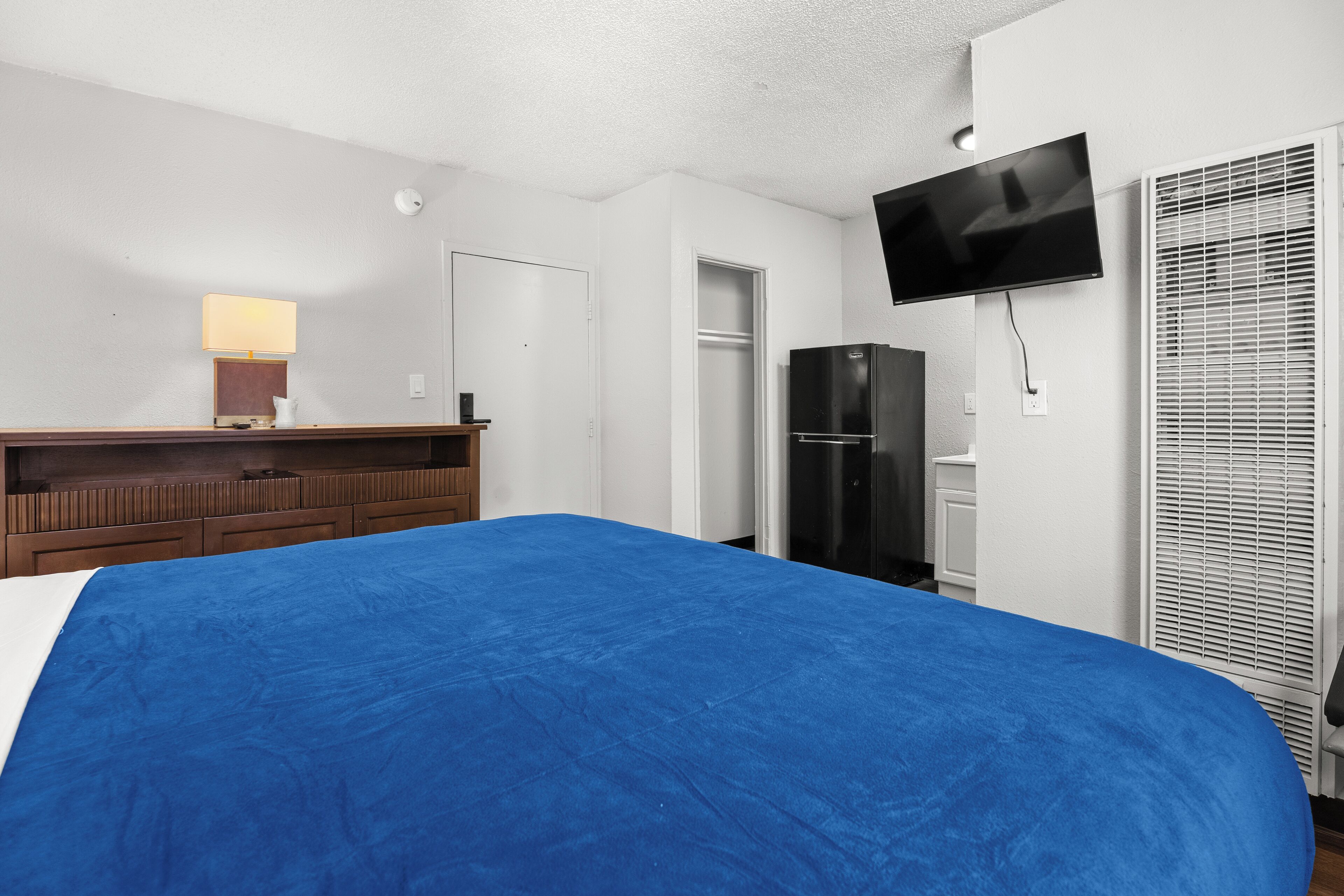 A hotel room with a large bed, a TV mounted on the wall, a refrigerator, a wooden dresser, and a window with blinds.