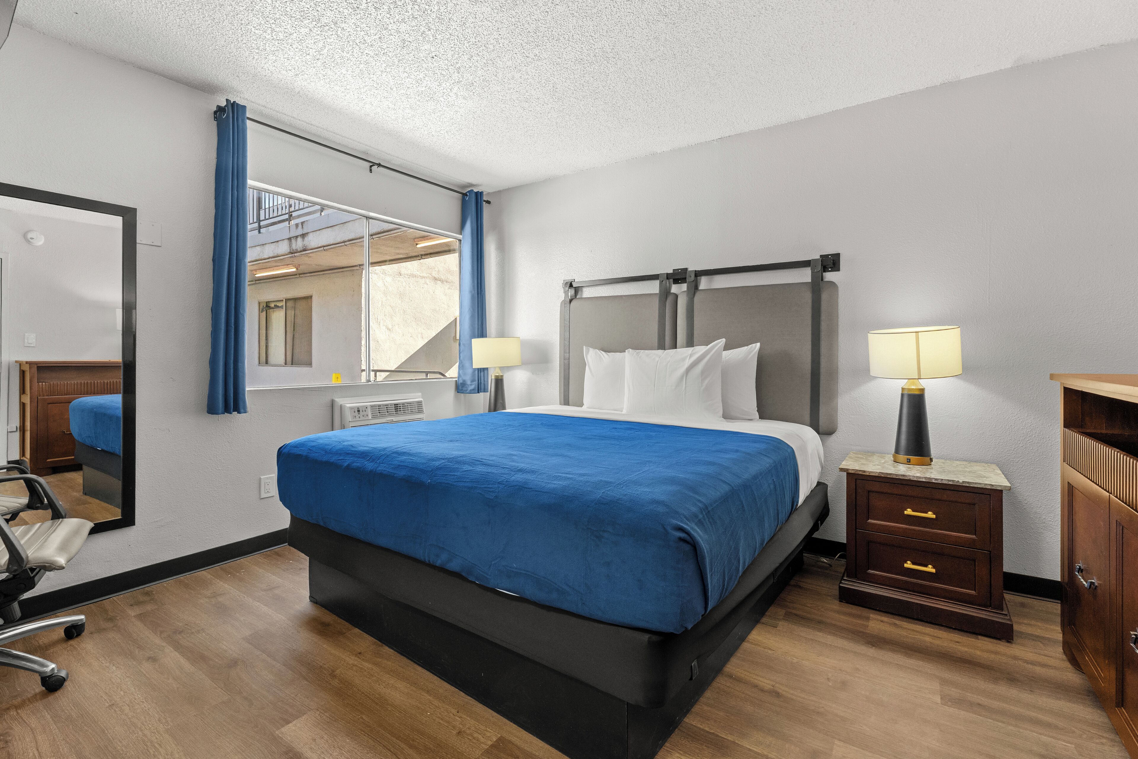 Une chambre d’hôtel avec un lit, une table de chevet, un miroir, une commode et une fenêtre avec des rideaux.