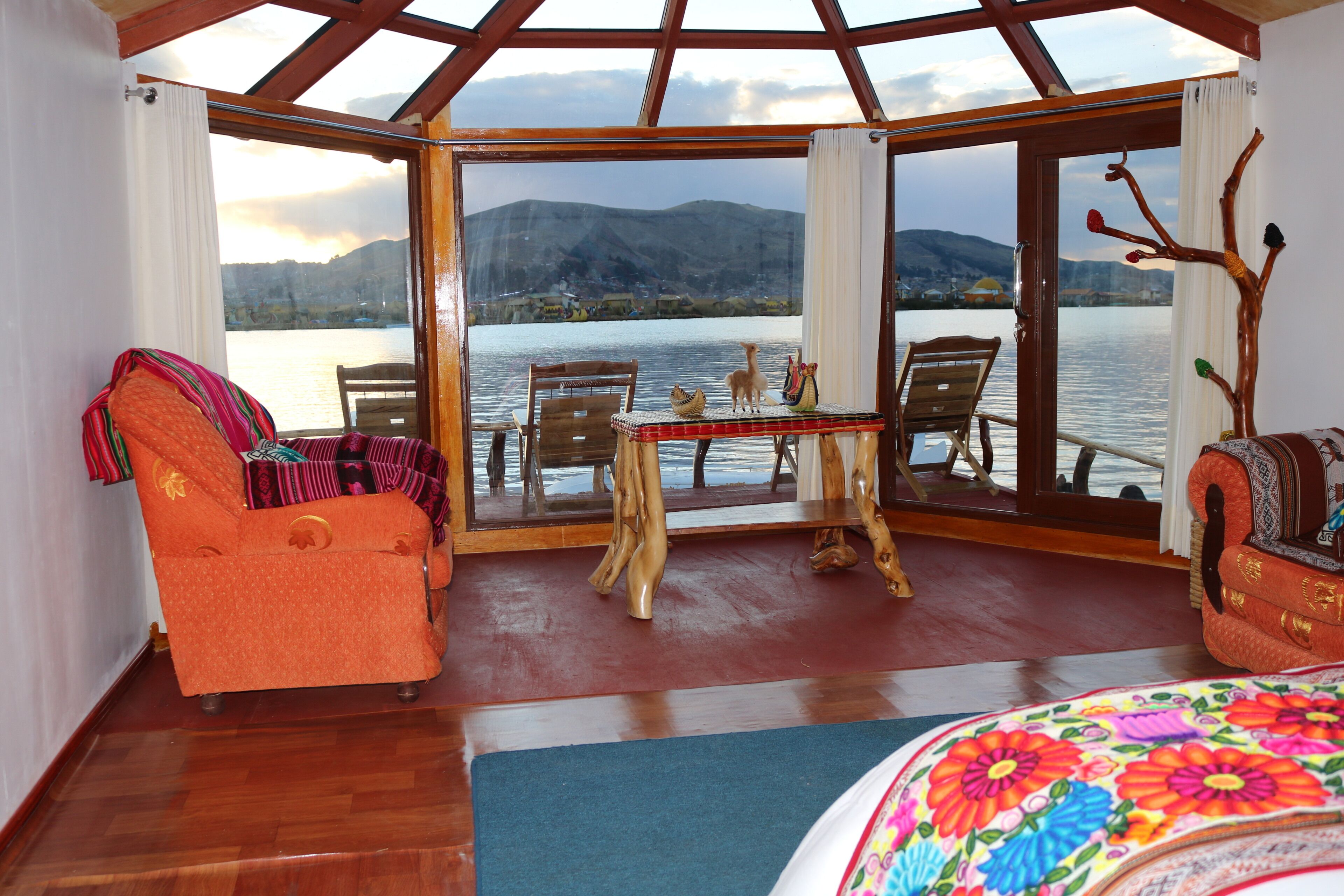A room with a wooden floor, a table with chairs, a sofa, and a view of a lake and mountains through large windows.