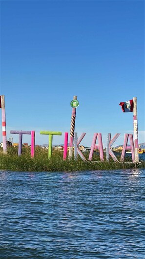 A sign with the word 'TIKARA' spelled out in colorful letters, with a flag on top, set against a clear blue sky and water.