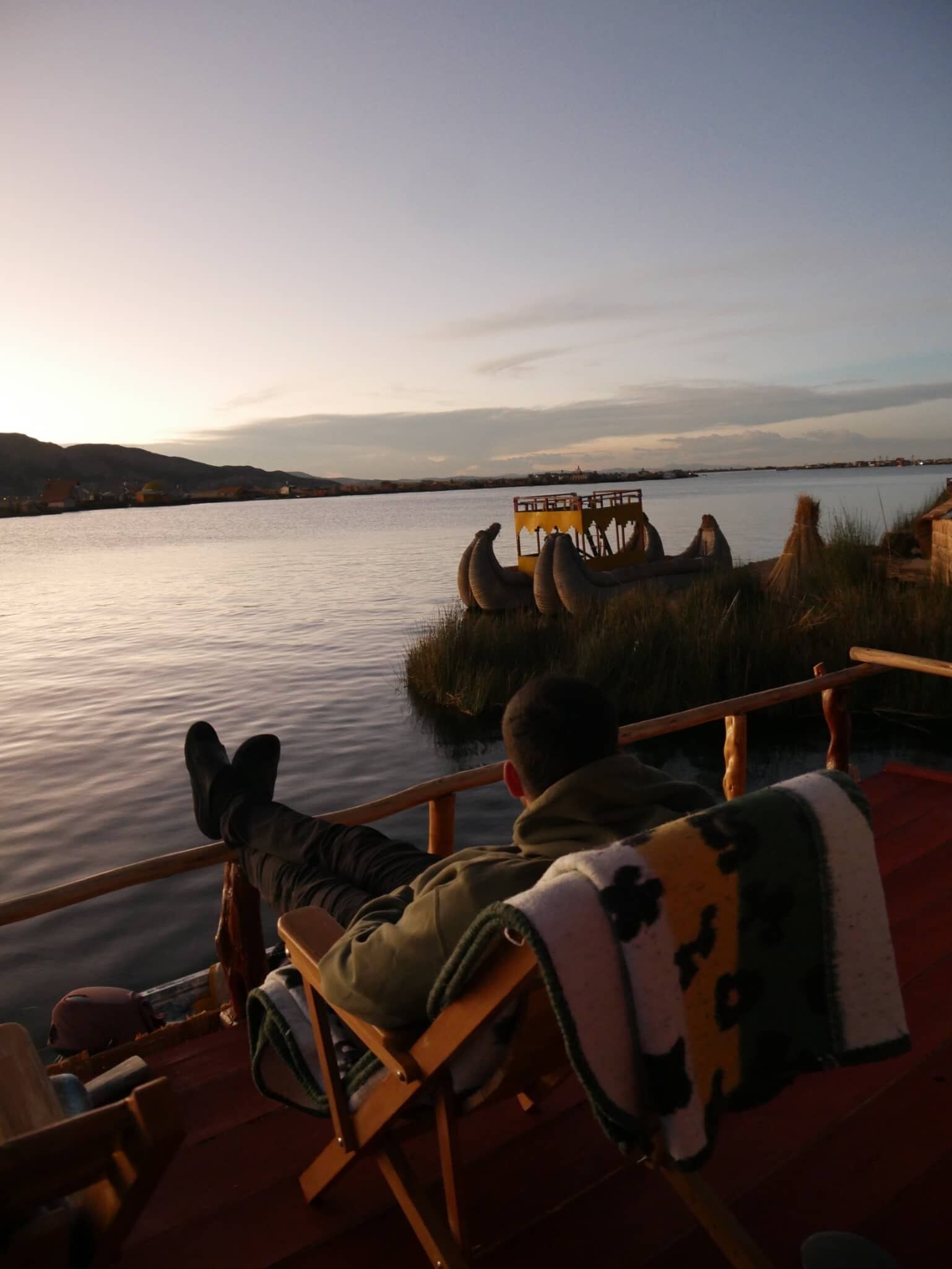 A person reclining on a deck chair overlooking a calm body of water with a distant shoreline and hills.