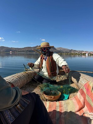 A person seated on a boat with a woven basket and a pot, surrounded by calm water and distant hills. - Little house on the floating islands of the Uros, Lake Titicaca, Puno-Peru (Puno)