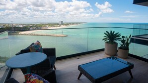 A balcony with a view of a beach and ocean, furnished with wicker chairs and a glass table. - Breathtaking 2BR deluxe condo with stunning views (Progreso)