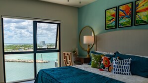 A bedroom with a large window offering a view of the sea and buildings, a bed with teal bedding, a desk with a lamp, and three colorful paintings on the wall. - Breathtaking 2BR deluxe condo with stunning views (Progreso)