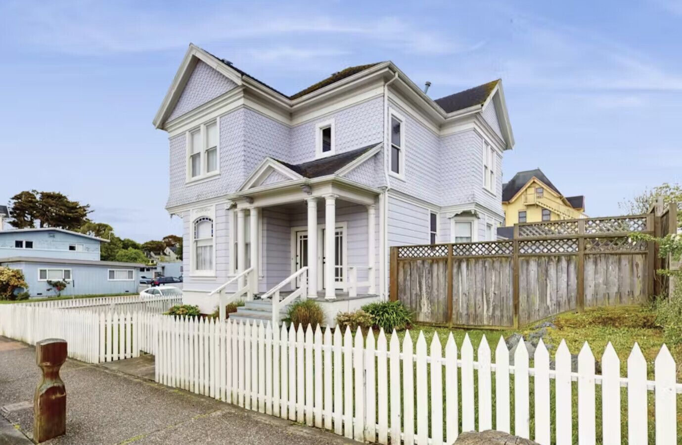A two-story house with a front porch, white picket fence, and a wooden fence in the backyard.