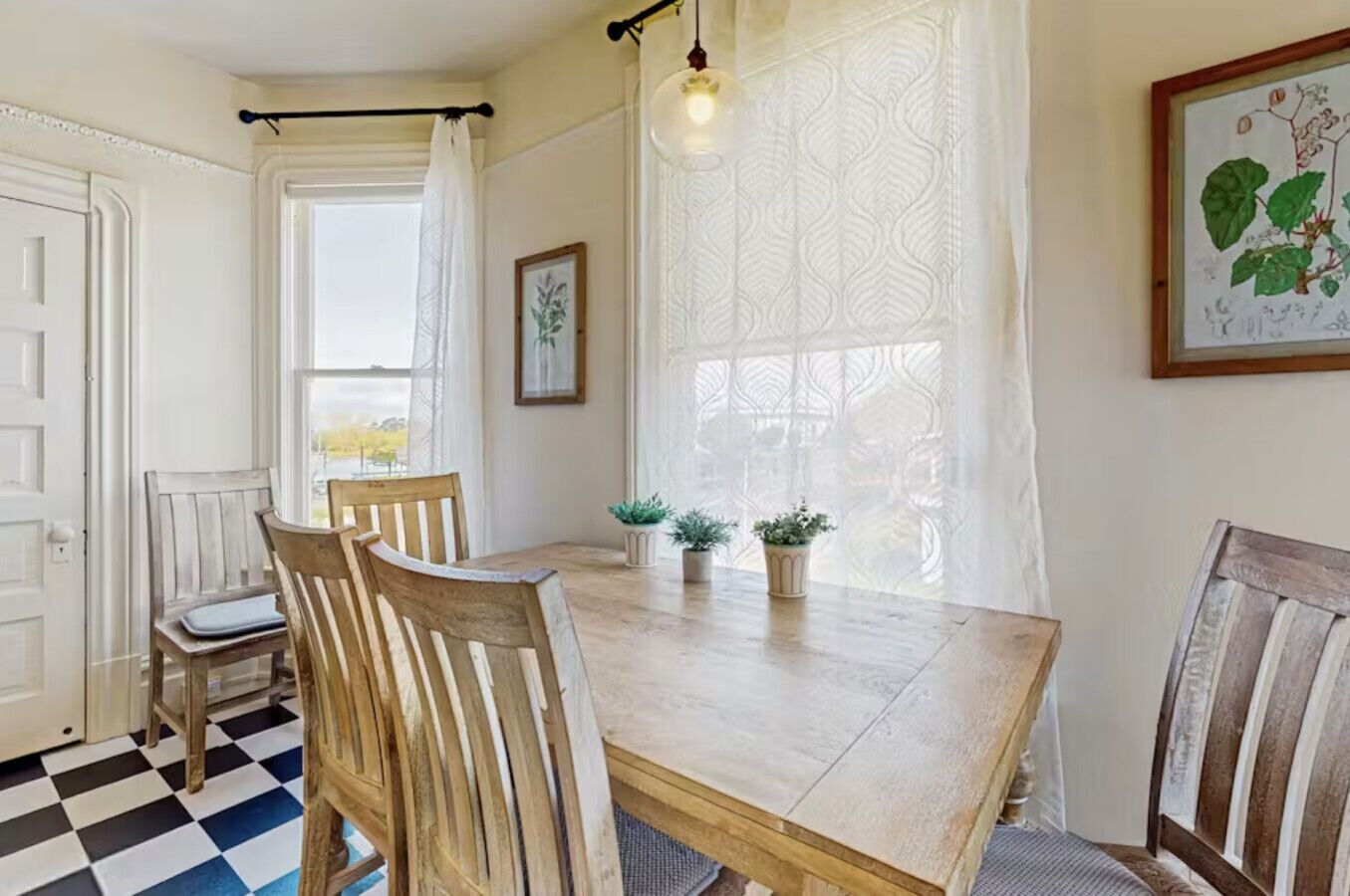 Un área de comedor con una mesa de madera, dos sillas, una planta en maceta y una ventana con cortinas estampadas.
