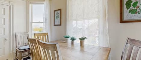 Un área de comedor con una mesa de madera, dos sillas, una planta en maceta y una ventana con cortinas estampadas.