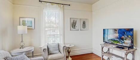 Un salon comprenant un canapé, un fauteuil, un téléviseur posé sur un meuble en bois, une lampe et, au mur, des reproductions encadrées de motifs botaniques.