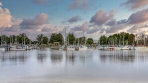 Een jachthaven met meerdere zeilboten afgemeerd, kalm water en bomen op de achtergrond.