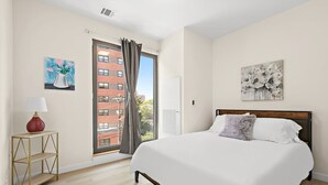 A bedroom with a bed, a nightstand with a lamp, a window with curtains, and artwork on the walls.