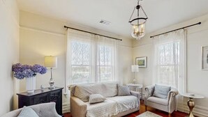 Un salon bien éclairé, avec un canapé beige, un fauteuil gris et un fauteuil blanc. On y trouve une petite table ronde avec une lampe, un vase décoratif avec des fleurs et un lustre.