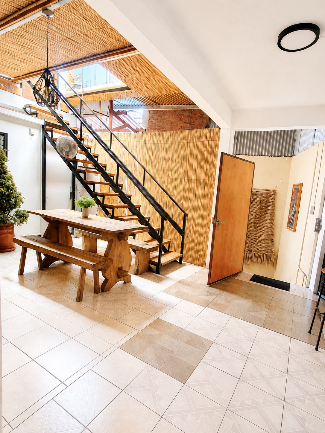 A spacious room with a wooden staircase, a dining table with benches, and a door leading to another area.