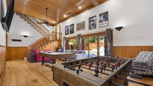 Amplia sala de estar con piso de madera, mesa de fútbol de mesa, mesa de billar y una escalera que lleva a un nivel superior.