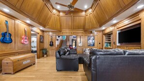 Un salón amplio con paneles de madera, un ventilador de techo, un televisor grande y varias guitarras colgadas en las paredes.