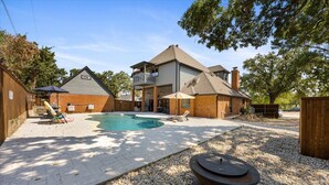 Una casa de dos pisos con un patio trasero que tiene una piscina, un hogar para fogata y un área de patio con mobiliario exterior.