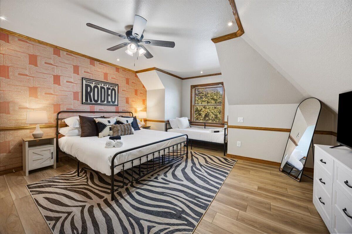 Une chambre à coucher comprenant un lit, un ventilateur de plafond, un tapis à motifs zébrés, une table de chevet, une commode et une fenêtre donnant sur des arbres.