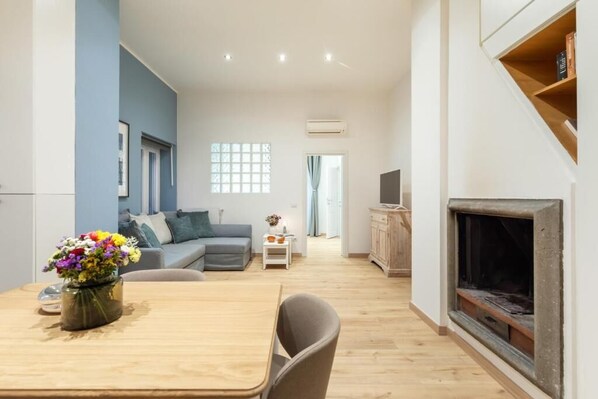A modern living room with a sofa, a dining table with chairs, a fireplace, and a TV on a wooden cabinet.