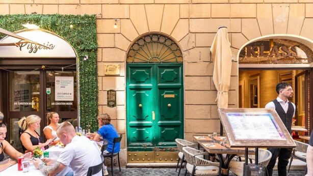 Un café al aire libre con mesas y sillas, una puerta verde y una pizarra con el menú.