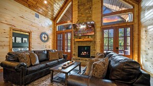 A cozy log cabin interior with leather sofas, a stone fireplace, and large windows.
