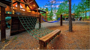 A wooden cabin with a rope swing and a hammock, surrounded by trees and a gravel path.
