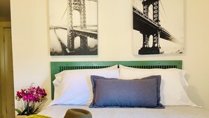 A hotel room with a bed, a green headboard, a decorative hat, and two black-and-white bridge paintings on the wall.