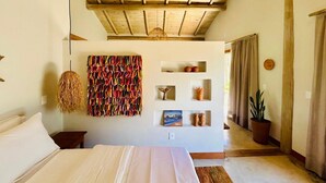 A bedroom with a wooden ceiling, a bed with white linens, a wall-mounted shelf with decorative items, and a hanging woven light fixture. - 🌴 Casa Preguiça in Corumbau – Comfort and Nature in Southern Bahia 🌊 (Prado)