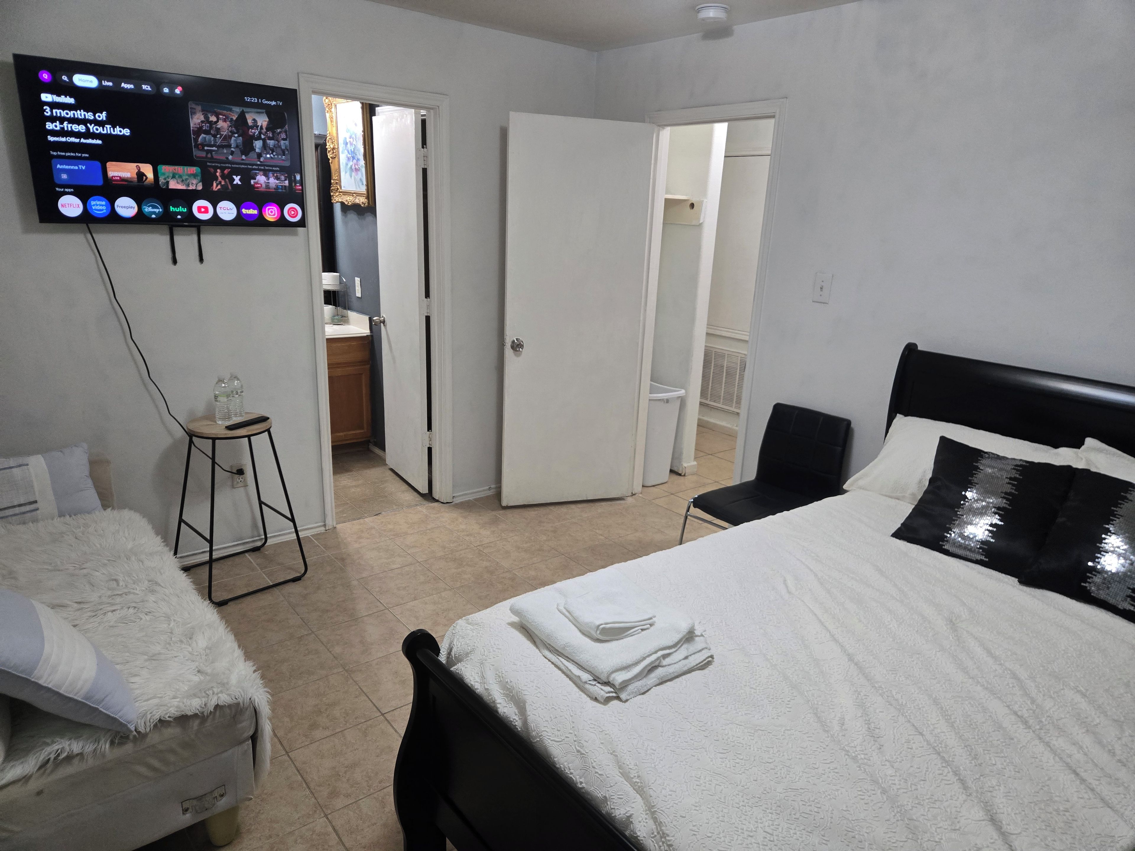 A hotel room with a bed, a chair, a small table, a TV displaying a YouTube channel, and a bathroom visible through an open door.