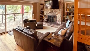 A spacious living room with a stone fireplace, leather sofas, a ceiling fan, and large windows.