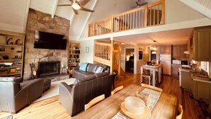 A spacious living area with a stone fireplace, leather sofas, a dining table, and a kitchen in the background.