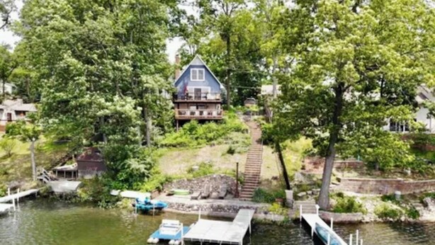 A house with a dock extending into a calm body of water, surrounded by lush greenery and trees.